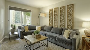 A living room with a grey sectional sofa, a wooden coffee table, and a floor lamp. There is a window with blinds, a small side table with a plant, and decorative wall panels. - Sandcastle at Avila a Family Condo, Steps to Beach (Avila Beach)