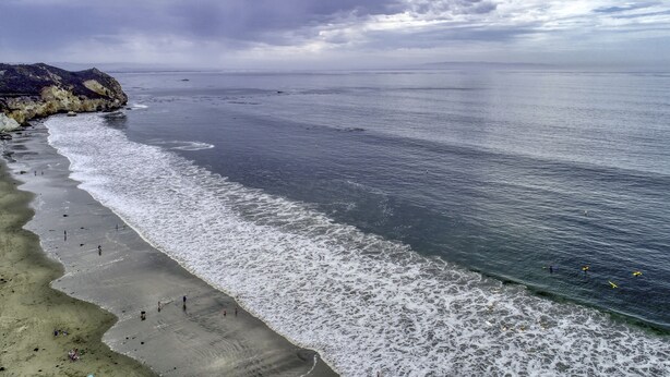 A coastal beach with waves gently lapping onto the shore, a rocky cliff on the left, and a clear view of the ocean. - Sandcastle at Avila a Family Condo, Steps to Beach (Avila Beach)