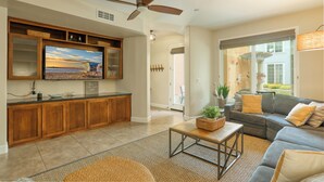 A spacious living area with a large flat-screen TV, wooden cabinets, a ceiling fan, a gray sofa with yellow cushions, and a view of the outdoors through a sliding glass door. - Sandcastle at Avila a Family Condo, Steps to Beach (Avila Beach)