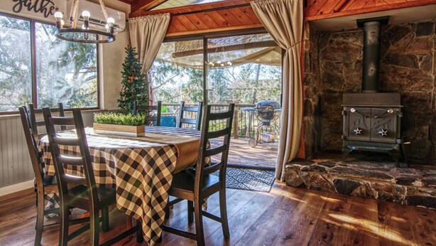 Un comedor acogedor con mantel a cuadros, sillas de madera y una araña. Se ve una chimenea de piedra, y un reloj está colgado en la pared.