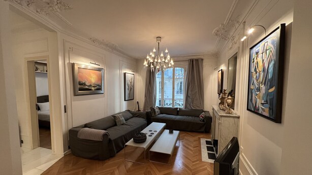 Ein modernes Wohnzimmer mit einer Couch, einem Couchtisch und einem Fernseher. An den Wänden hängen Gemälde und es gibt eine Deckenleuchte.