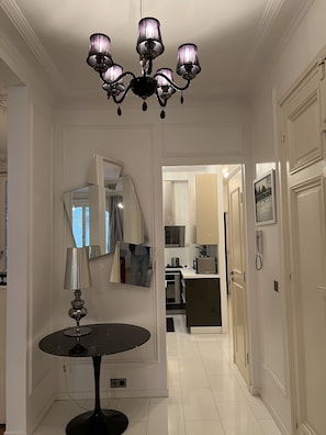 A hallway with a chandelier, a round table with a lamp, a mirror, and a kitchen area in the background.