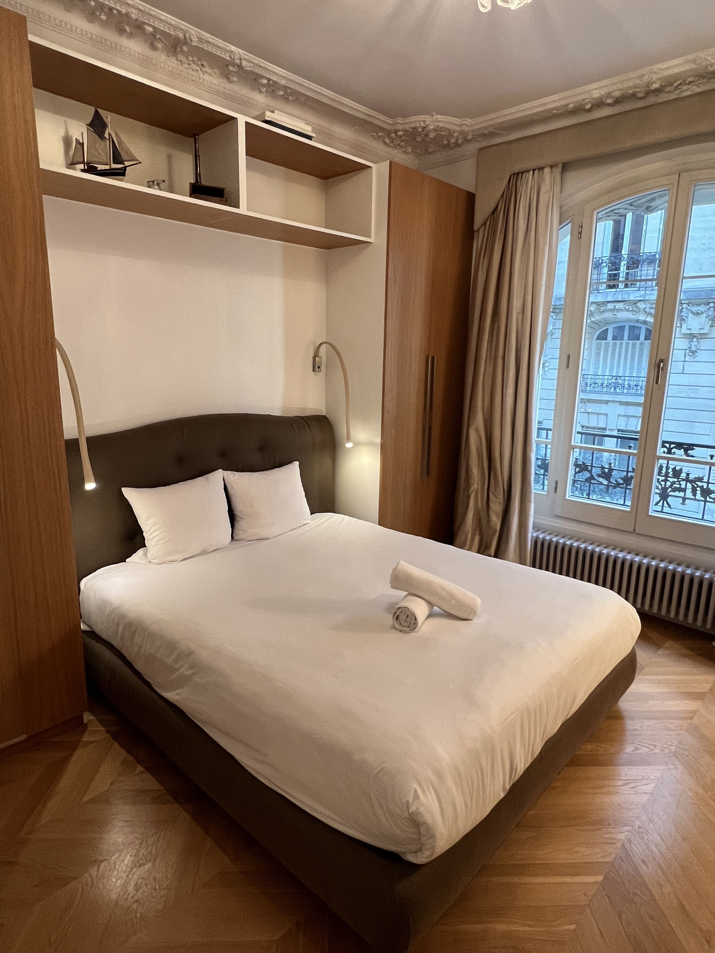Ein ordentlich bezogenes Bett mit einem zusammengerollten Handtuch, einem Kopfteil und einem Nachttisch mit einer Lampe.