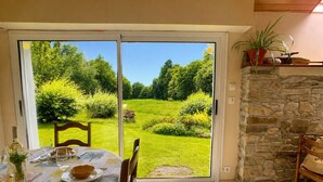 A dining area with a table set for two, a stone wall, and a view of a green garden through a sliding door. - TY AR STIVELL, the balance between land and sea 4-7PERS, MOELAN / SEA, WIFI, BEACHES (Moëlan-sur-Mer)