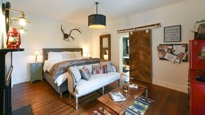 A bedroom with a bed, bedside lamps, a hanging light fixture, a wooden floor, a sofa, a coffee table, and a red appliance.