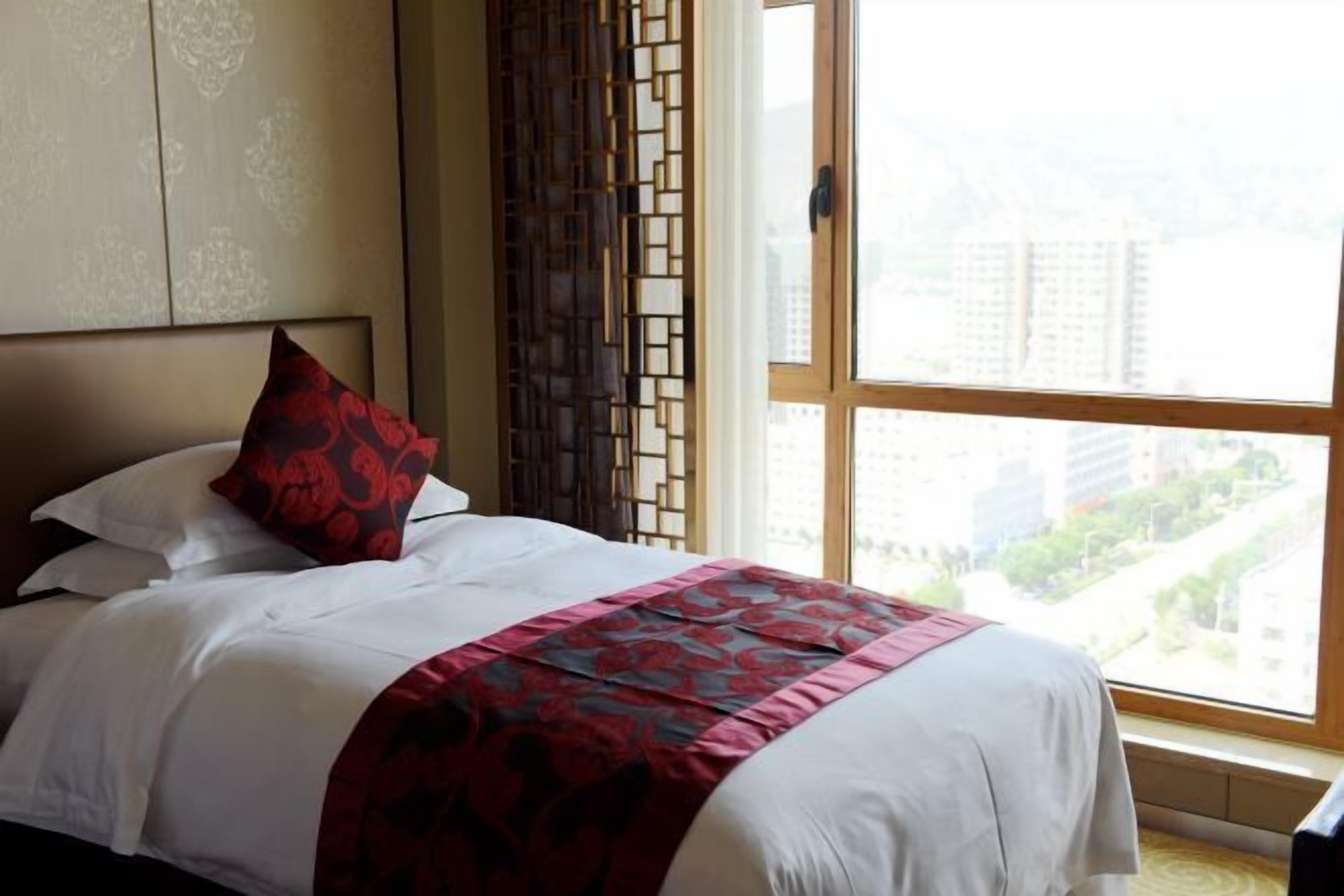 A hotel room with a bed, a window with a view of buildings, and a decorative screen.