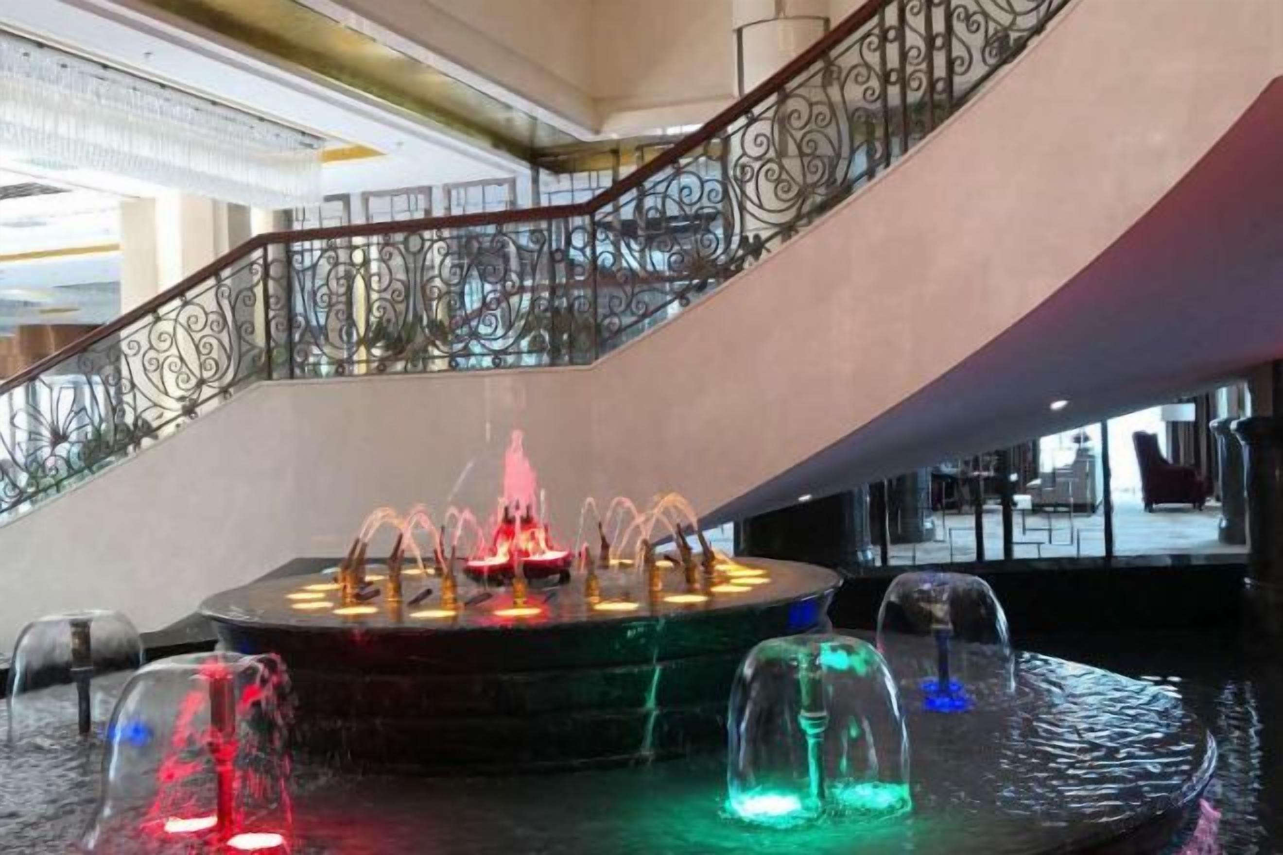 Un espace intérieur à plusieurs niveaux, doté d’un élément aquatique central, d’un escalier orné de balustrades travaillées et d’une surface réfléchissante au niveau inférieur.