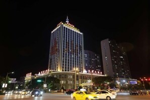 Un hotel muy iluminado con una estructura vertical destacada y un techo plano, rodeado por una calle concurrida y vehículos.