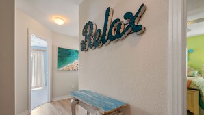 En gang med en træbænk, et skilt med teksten 'Relax', et maleri af en strand og en dør, der fører til et værelse med en seng.