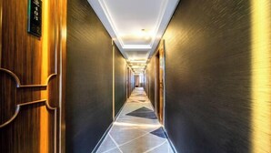 Un largo y estrecho pasillo de hotel con un suelo de patrón geométrico, puertas de madera en un lateral y una lámpara moderna en el techo.