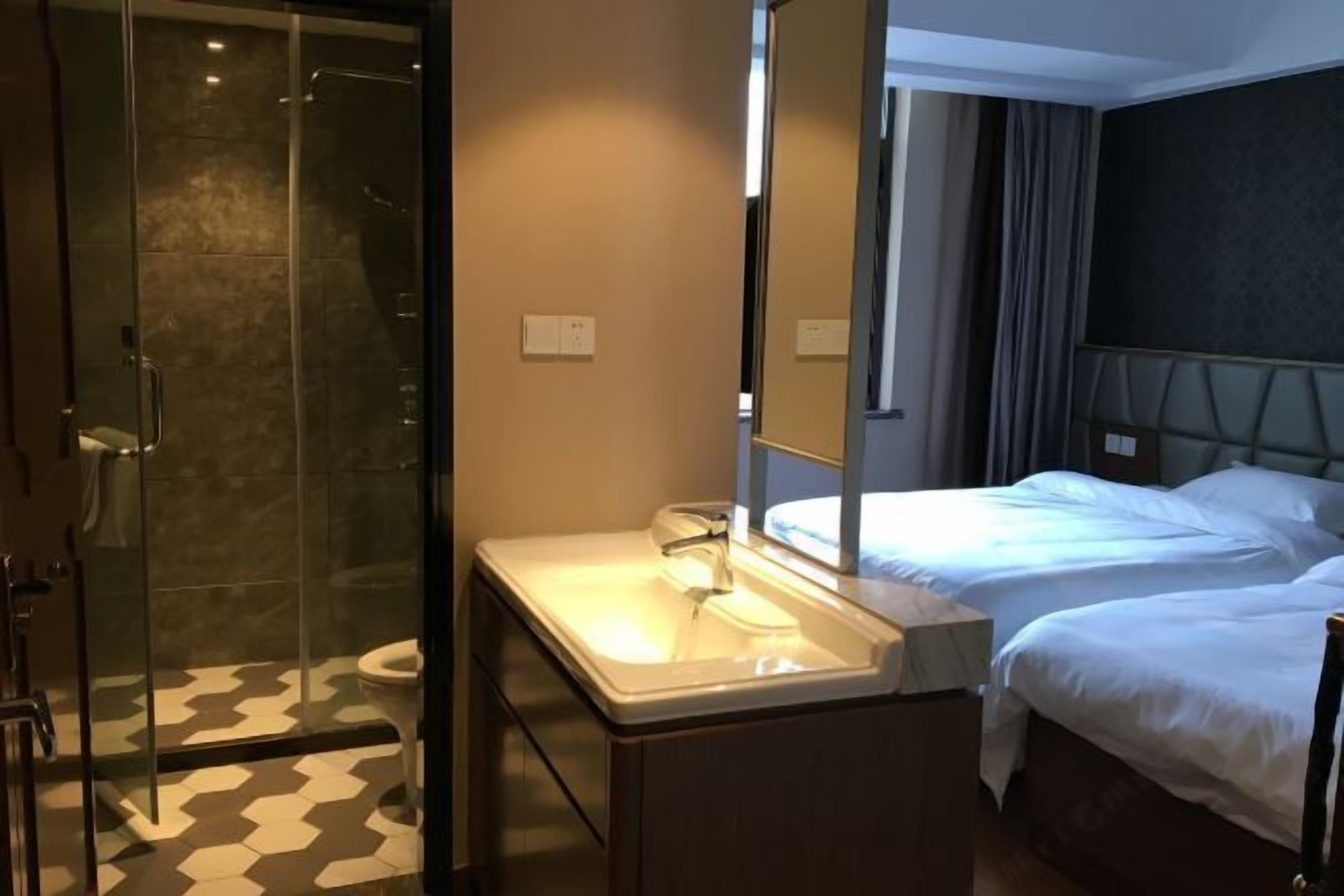 Habitación de hotel con ducha envuelta en vidrio, lavamanos blanco, dos camas y un espejo grande.