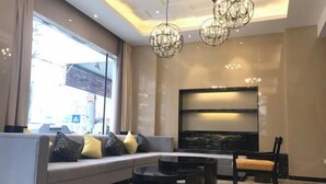 Un salón moderno con un sofá gris, una mesa de centro negra y un televisor de pantalla plana grande. Hay dos candelabros de cristal y una ventana que proporciona luz natural.