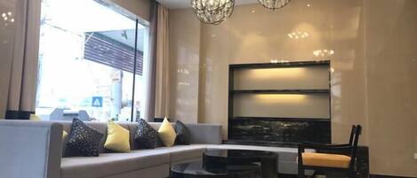 Un salón moderno con un sofá gris, una mesa de centro negra y un televisor de pantalla plana grande. Hay dos candelabros de cristal y una ventana que proporciona luz natural.