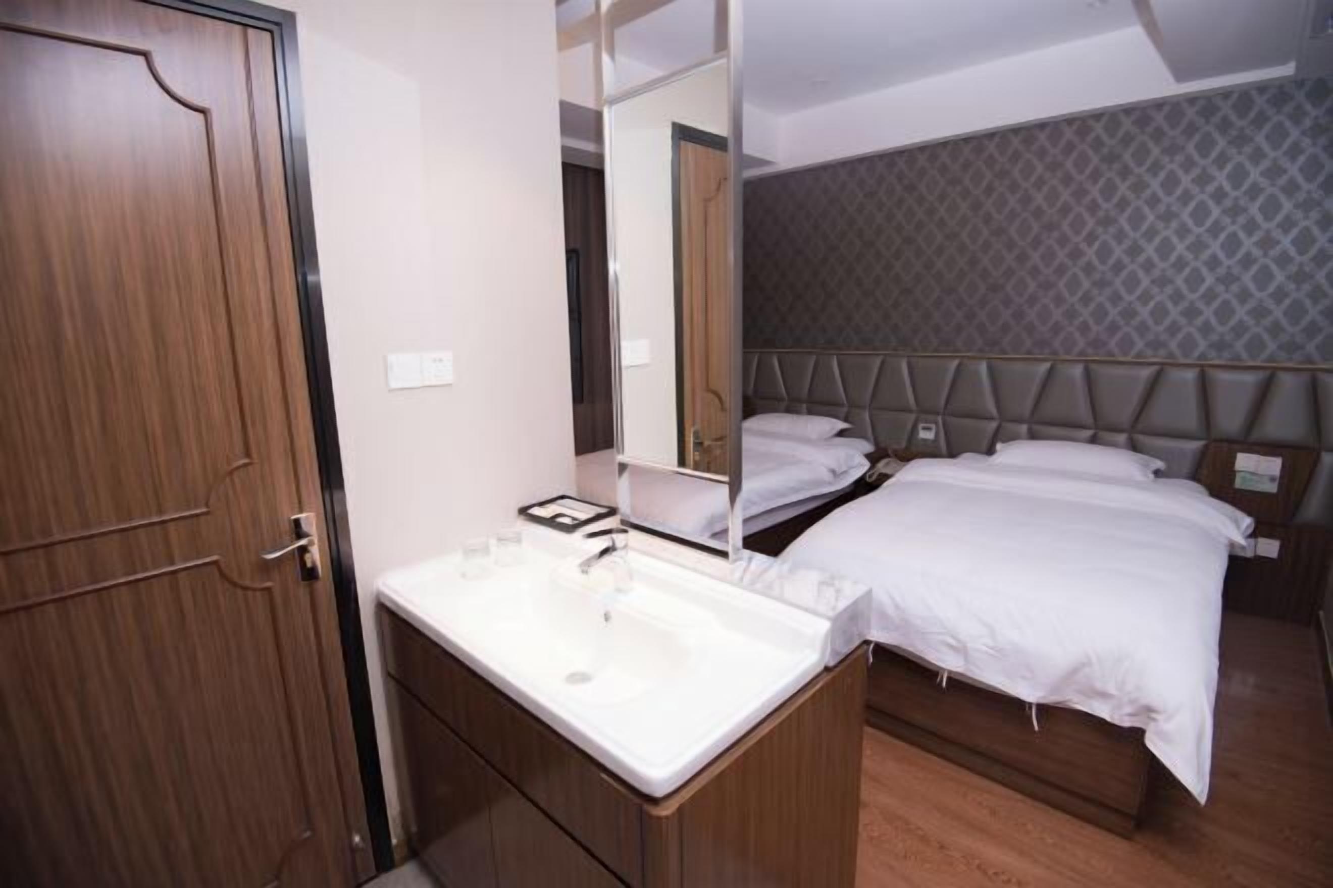 A hotel room with a wooden door, a white sink, a mirror, a bed with a headboard, and a patterned wall.