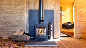 A wood-burning stove with logs, a basket of firewood, and a bed in a wooden room. - The Safari Lodge at Rusty Lane Farm (Seend)