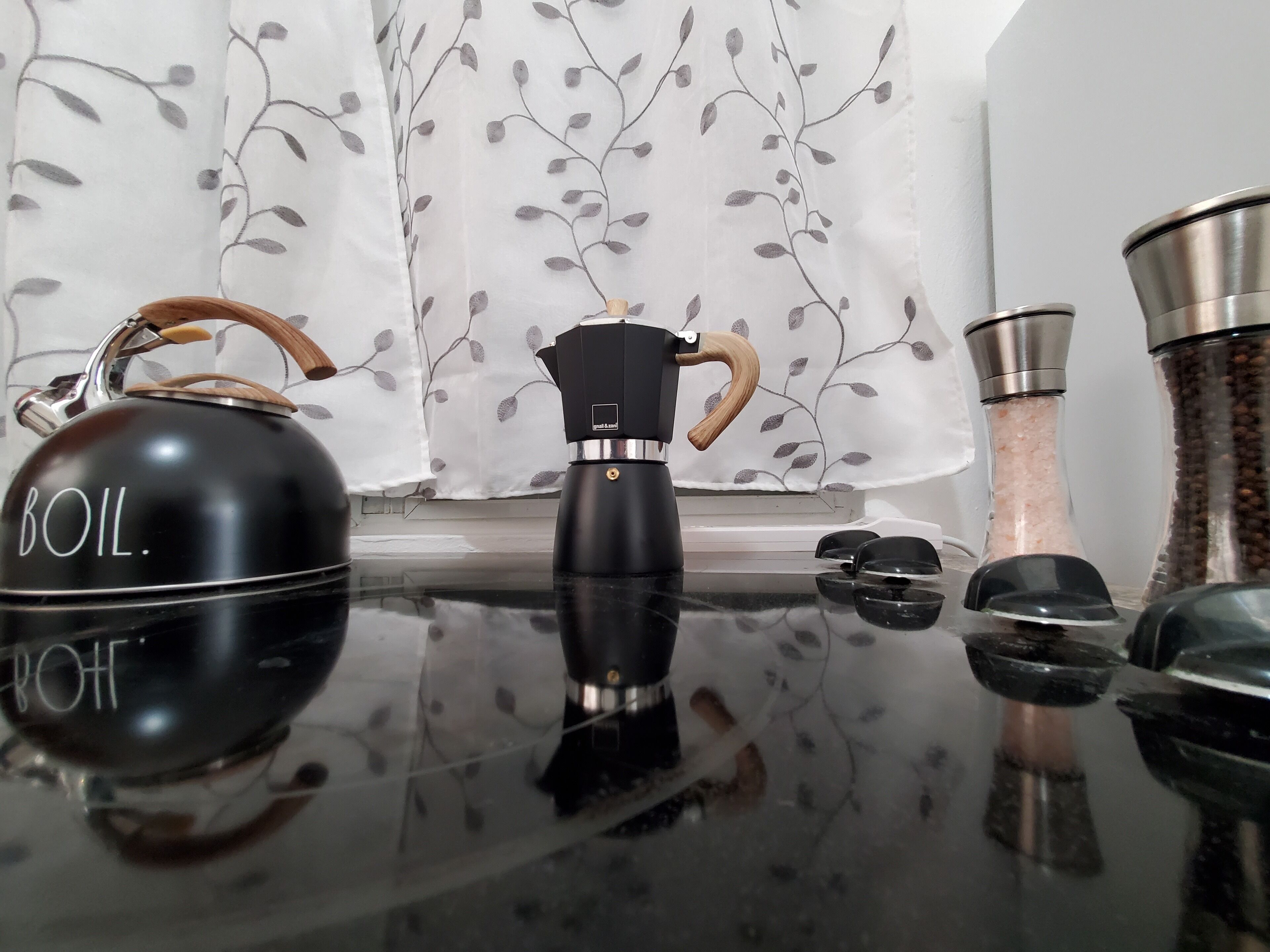 Une cuisine avec une bouilloire noire portant l’inscription « À FAIRE BOUILLIR », une machine à café et un salière posés sur un plan de travail réfléchissant.
