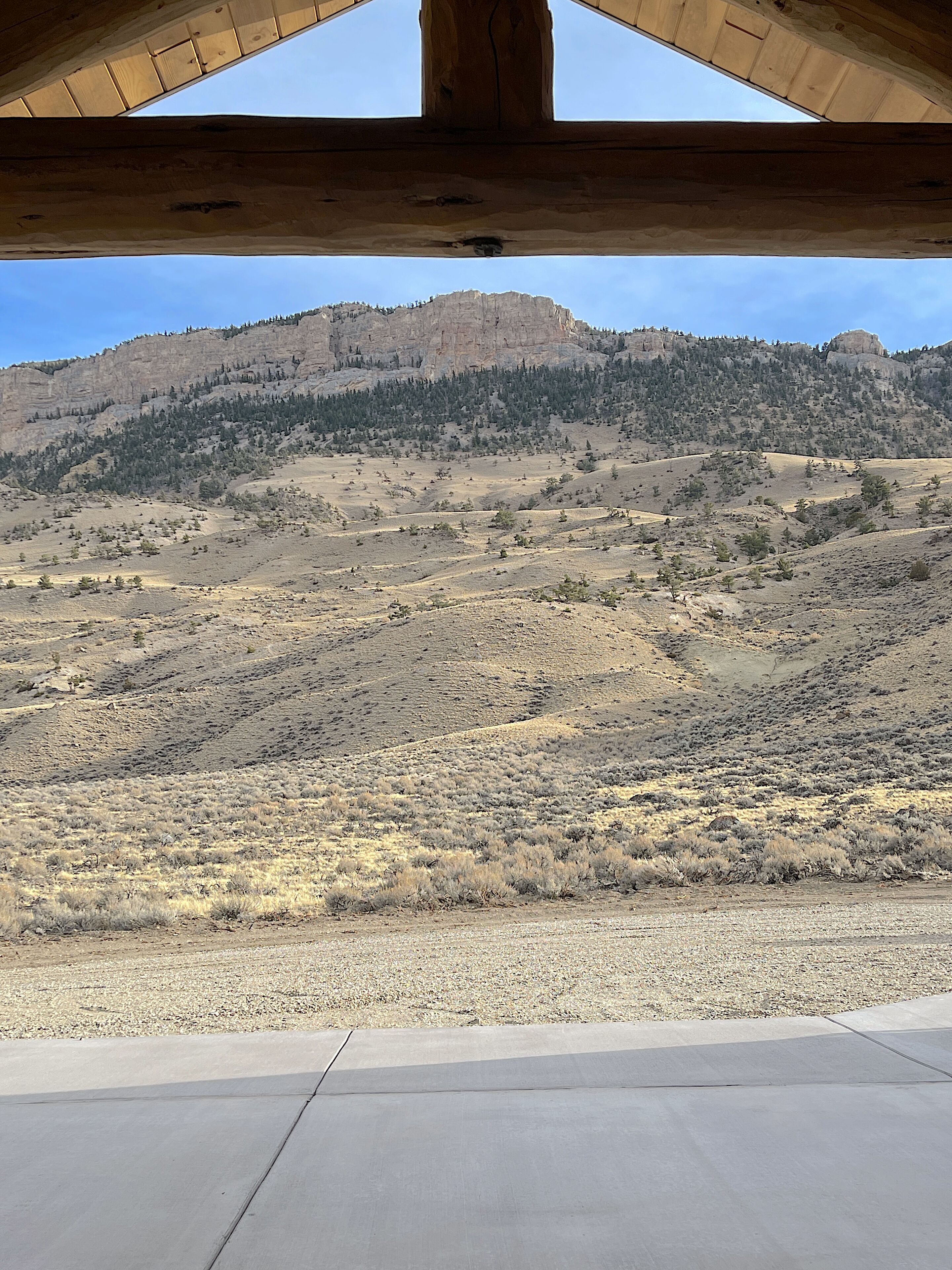 Vista desde el interior de un edificio con suelo de concreto y vigas de madera, que da a un paisaje seco y montañoso con vegetación escasa y una montaña boscosa a lo lejos.