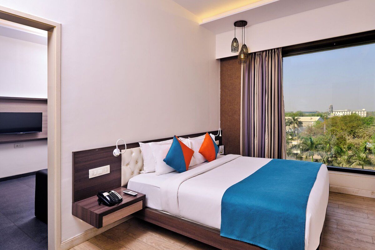 A modern hotel room with a large bed, a bedside table with a phone and a lamp, a TV on the wall, and a view of the city through large windows.
