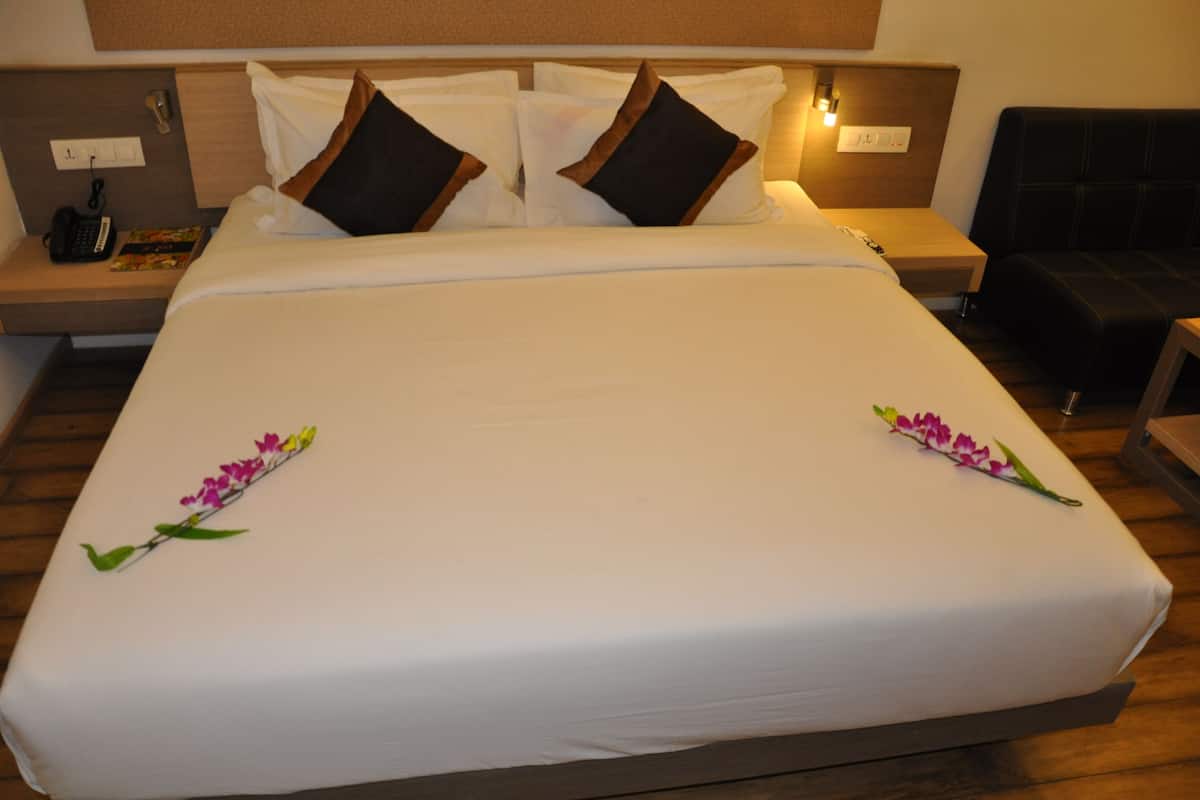 A neatly made bed with white linens and brown pillows, a bedside table with a phone and a book, and a small nightstand with a lamp.