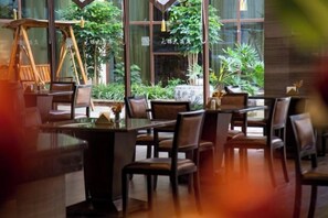 Un comedor interior con mesas y sillas, ventanas amplias y plantas en macetas.