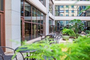 Un área de estar al aire libre con mesas y sillas, rodeada de plantas en macetas y un edificio con grandes ventanales.