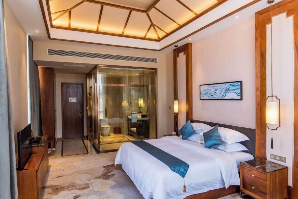 Una habitación de hotel con una cama grande, un baño con paredes de vidrio y una mesita de noche de madera.