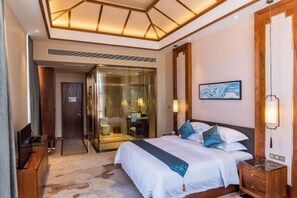 Una habitación de hotel con una cama grande, un baño con paredes de vidrio y una mesita de noche de madera.