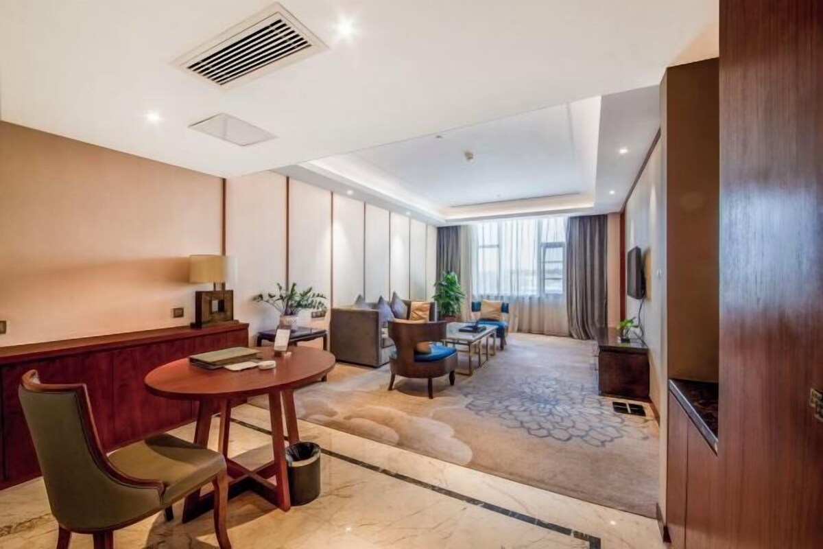 Una habitación de hotel con zona de comedor, un conjunto de sofás, un televisor y un ventanal grande.