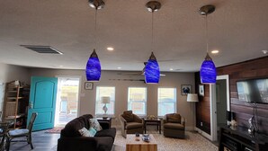 Uma sala de estar moderna com área de cozinha, incluindo uma ilha grande com pia, três luminárias pendentes e uma televisão.