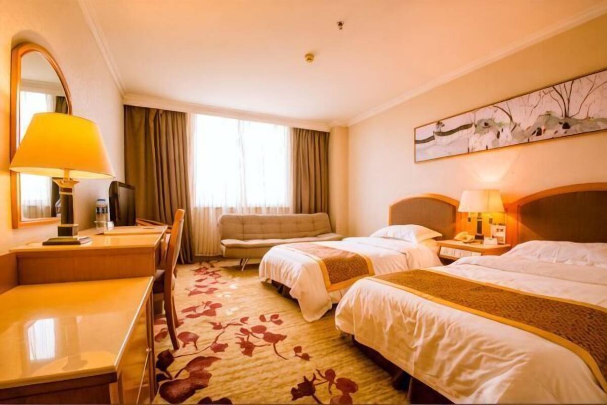 A hotel room with two beds, a desk, a chair, a sofa, a lamp, a painting, and a window with curtains.