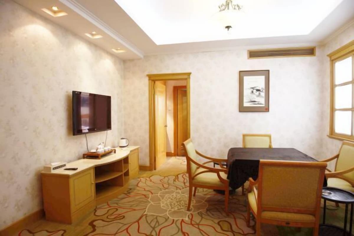 A hotel room with a flat-screen TV mounted on the wall, a dining table with chairs, a small cabinet, and a patterned carpet.
