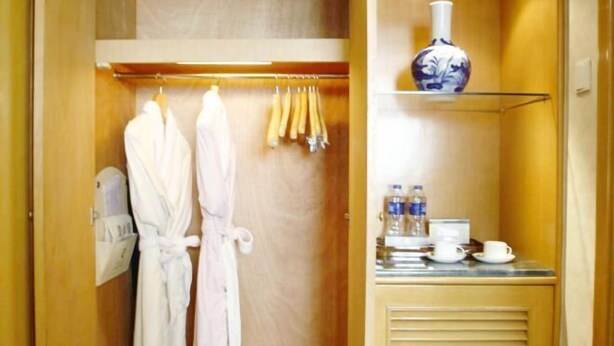 A hotel room with a wooden wardrobe containing two white robes, a hanging rack with towels, and a shelf with a vase, water bottles, and a cup. - Jinhu Hotel (Wuxi)