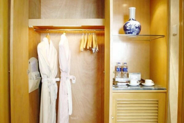 Habitación de hotel con un armario de madera que contiene dos albornoces blancos, un perchero con toallas y un estante con un florero, botellas de agua y una taza.