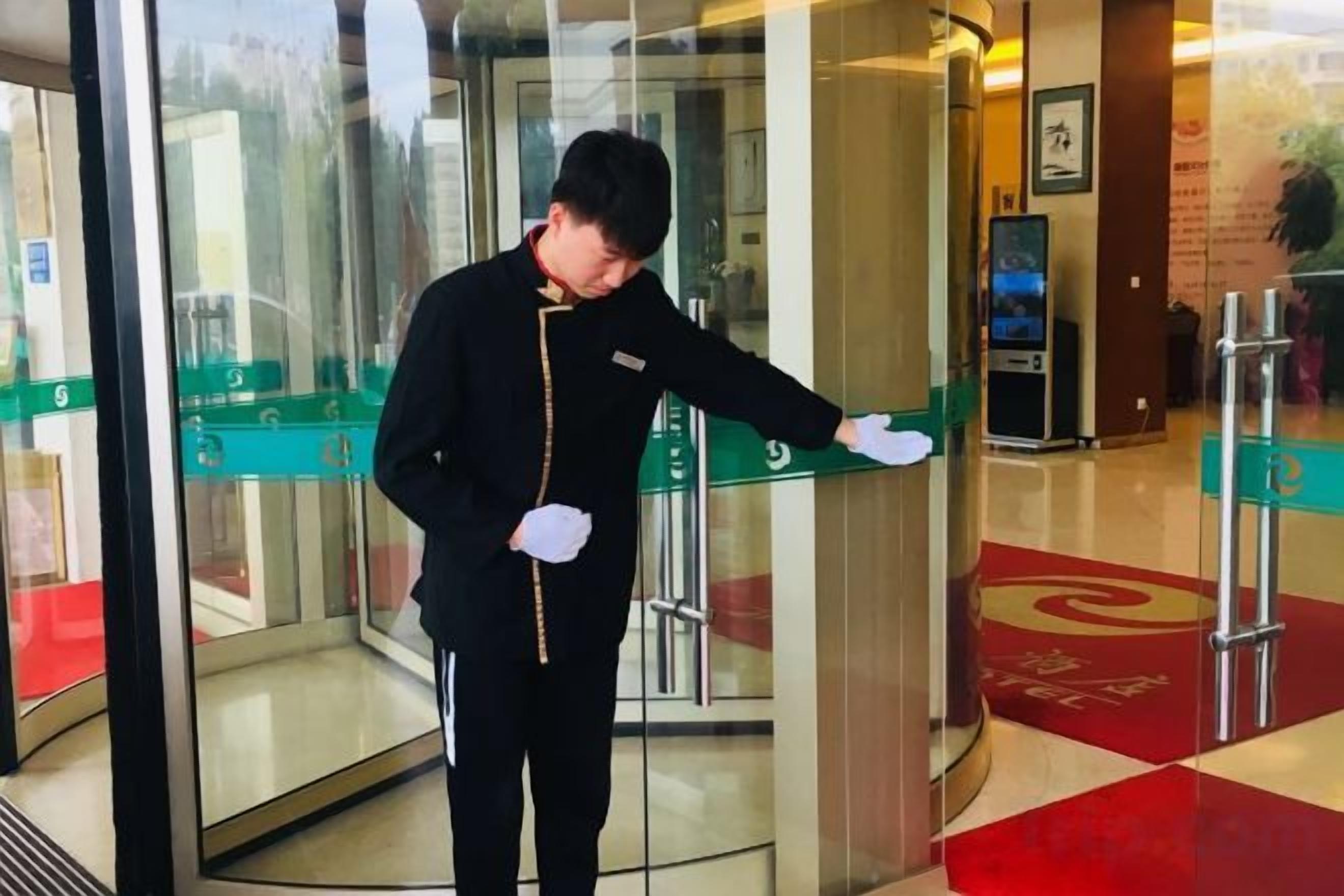 Una persona con uniforme de pie en una puerta giratoria, con un panel de vidrio y un suelo alfombrado de color rojo a la vista.