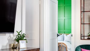 A room with a wooden entertainment center, a television, a green-lit bedroom, a teal armchair, and a colorful rug.