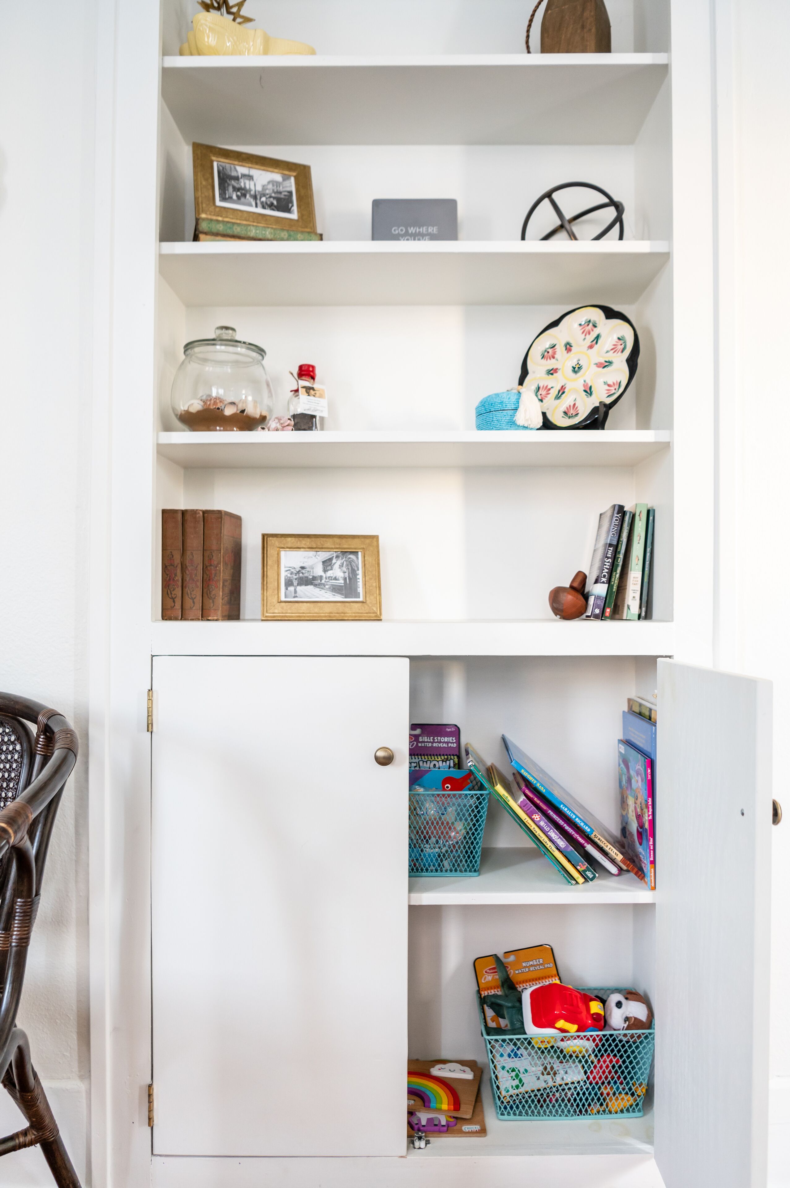 A white cabinet with shelves containing books, a framed photo, decorative items, and a wicker chair.