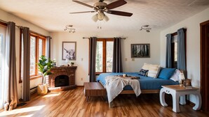 A cozy room with a fireplace, a bed with blue bedding, a wooden floor, and a ceiling fan. - Lakefront  Hot tub. Amazing views. 35 min to Indy (Martinsville)
