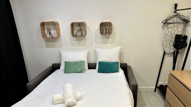A neatly made bed with white bedding and two green pillows, a wooden bedside table, and a wall-mounted shelf with decorative items. - Apartment Gutenberg 3 (Paris)