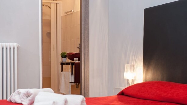 A hotel room with a red bed, white towels, a visible bathroom with a shower, and a nightstand with a lamp. - Domus Fabiola Vaticano Matrimoniale (Roma)
