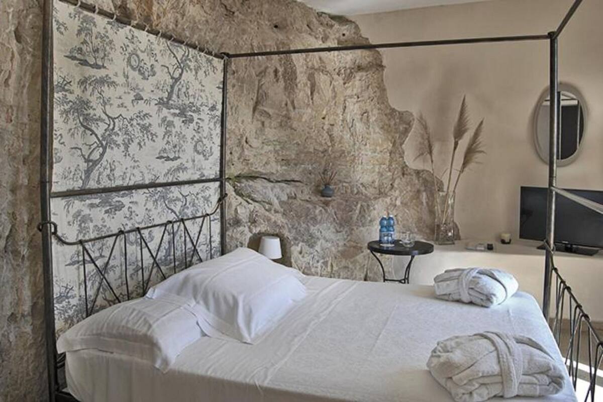 Een slaapkamer met een stenen muur, een bed met witte lakens, een klein tafeltje met een fles water, en een televisie op een standaard.