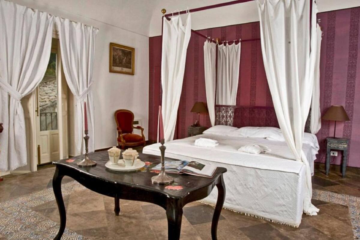 Een bed met hemelbed voorzien van witte lakens, een tafel met een dienblad en een rode stoel in een kamer met een kamerbreed tapijt en rode wanden.