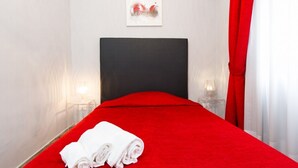 Ein Hotelzimmer mit roter Bettdecke, einem ordentlich gefalteten weißen Handtuch auf dem Bett, einem schwarzen Kopfteil, einem gerahmten Kunstwerk an der Wand und einem roten Vorhang.