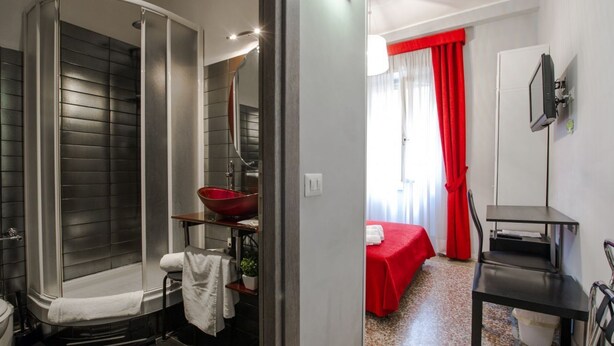 Ein Badezimmer mit Dusche und Waschbecken, im angrenzenden Raum ein rotes Bett.