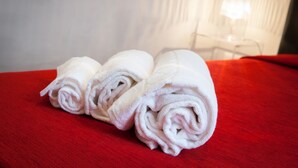 Drei aufgerollte Handtücher auf einer roten Bettdecke in einem Zimmer mit weißer Wand und einem Fenster mit Jalousien.