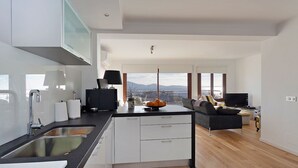 A modern kitchen with a central island, a dining area with a sofa and a TV, and large windows offering a view of the city. - Seafront Penthouse Marina Apartment (Port de Pollença)