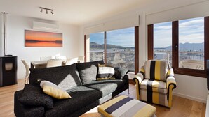 A modern living room with a large sofa, a striped armchair, and a view of a cityscape through large windows. - Seafront Penthouse Marina Apartment (Port de Pollença)