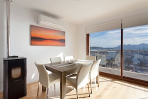 A modern dining area with a table and chairs, a large window with a marina view, and an air conditioning unit on the wall. - Seafront Penthouse Marina Apartment (Port de Pollença)