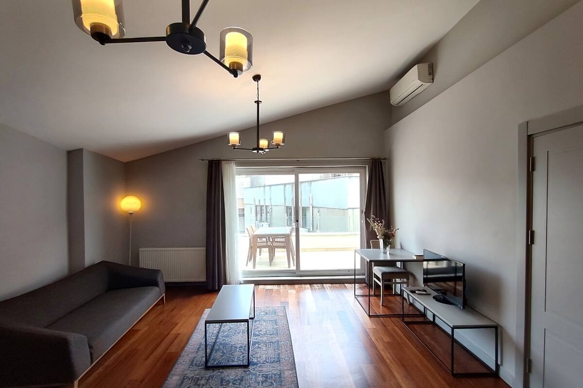 A modern living room with a sofa, a coffee table, a dining table with chairs, and a large sliding glass door leading to an outdoor area.