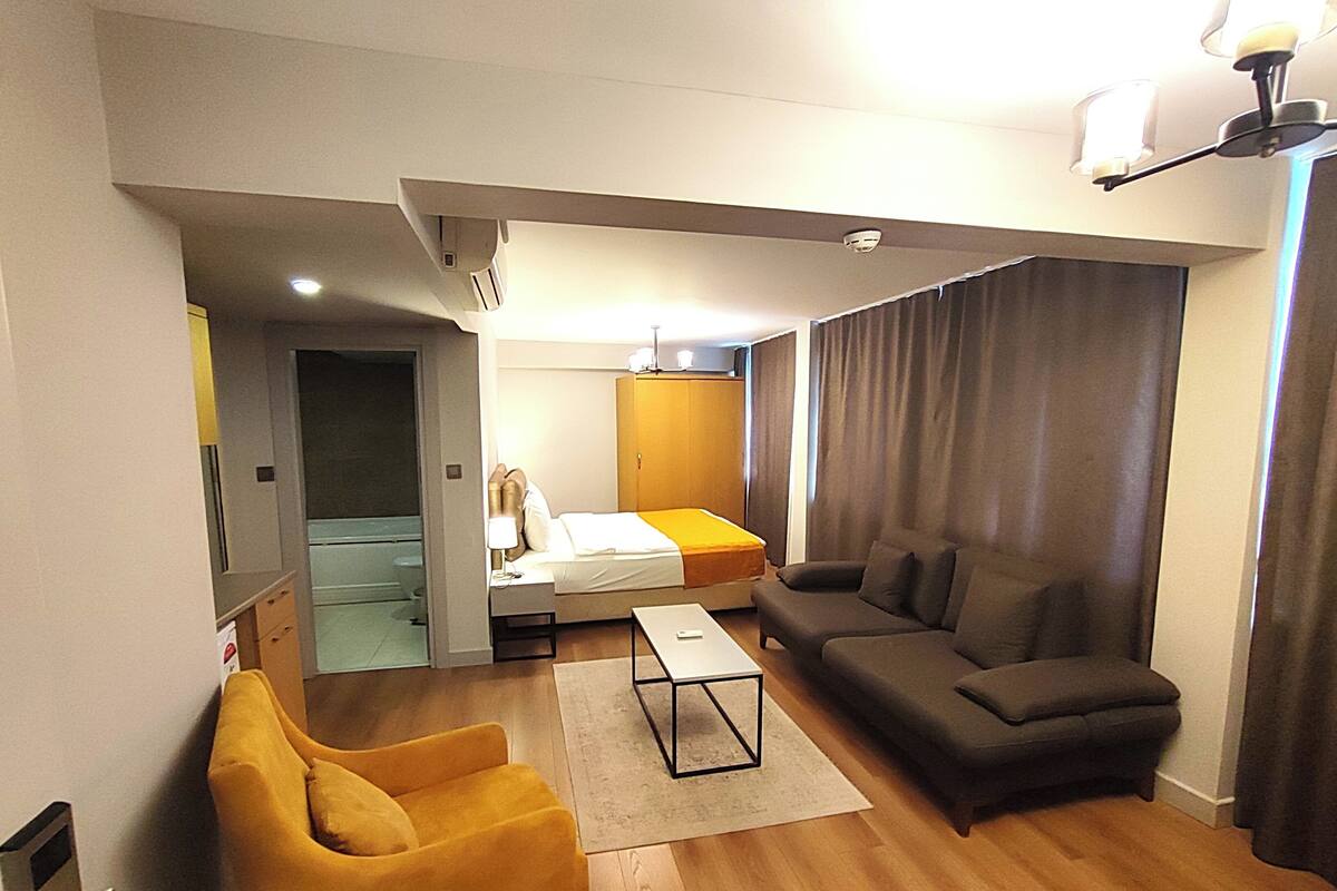 A compact hotel room with a bed, a sofa, a yellow armchair, and a small table.