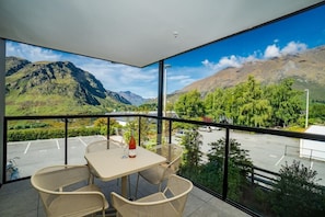 A balcony with a table and chairs, a bottle of wine, and a view of mountains and a parking lot.
